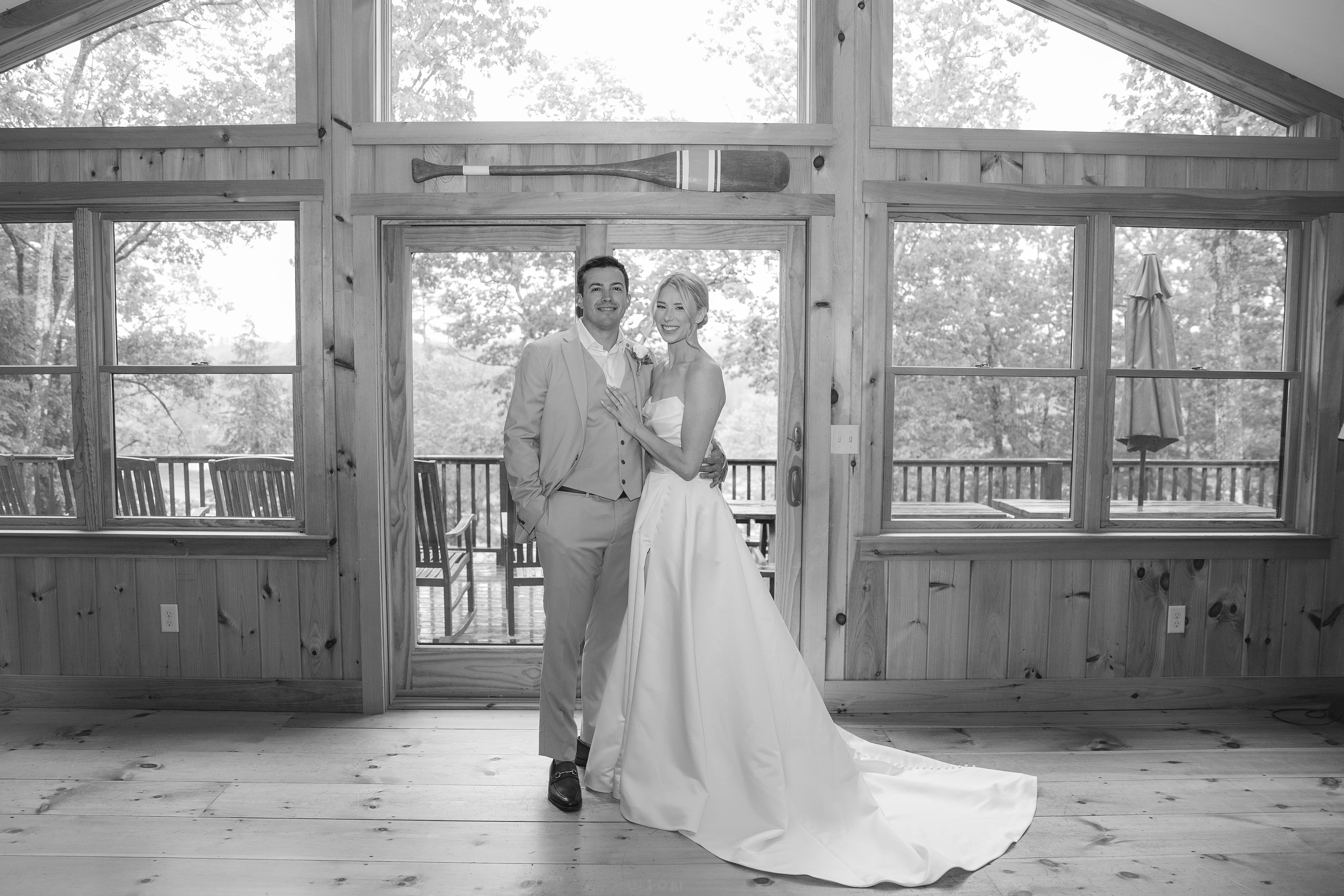 A bride and groom pose together in a wooden cabin with large windows overlooking a natural setting.