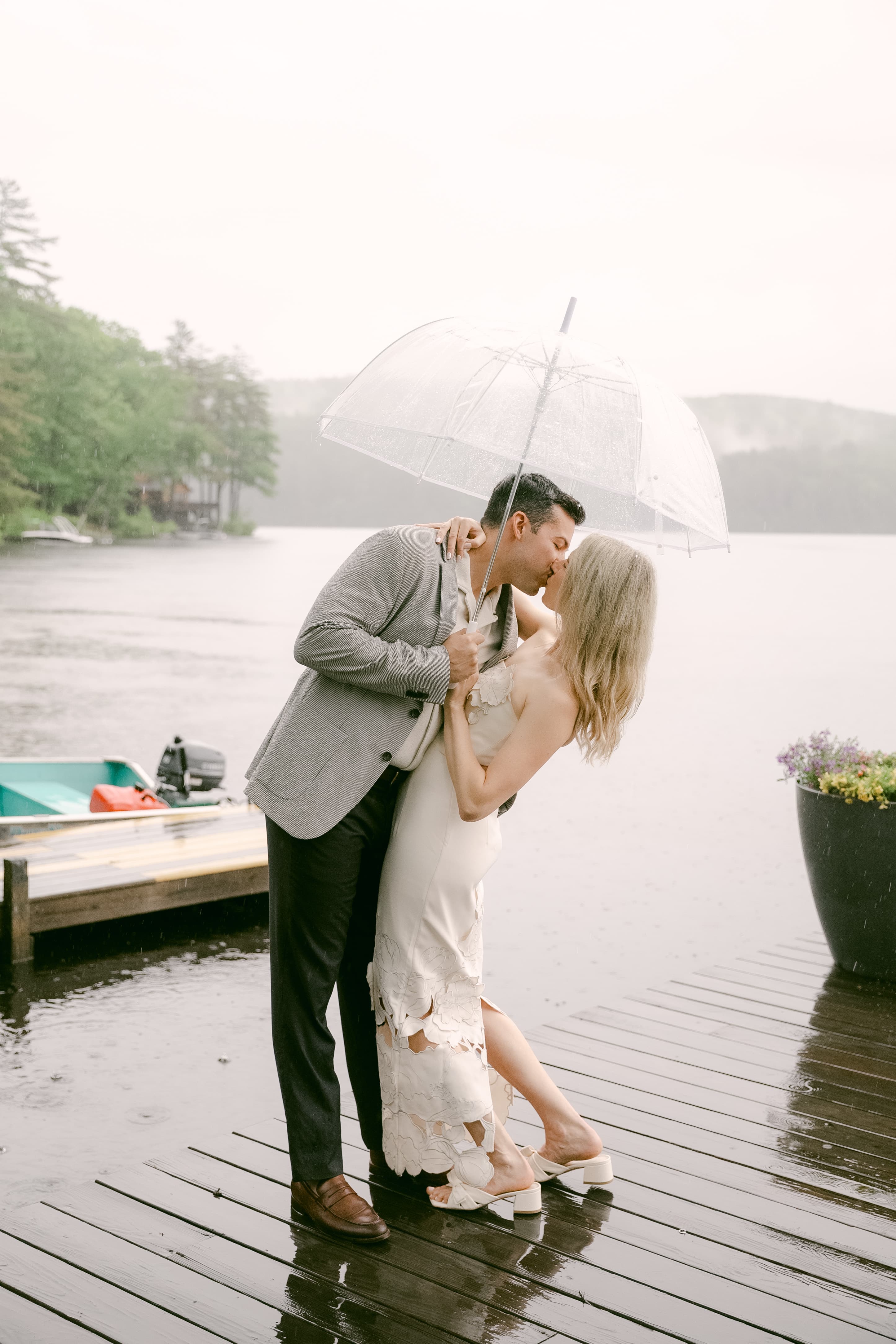 A couple kisses under a clear umbrella while standing on a wet dock by a lake in the rain.