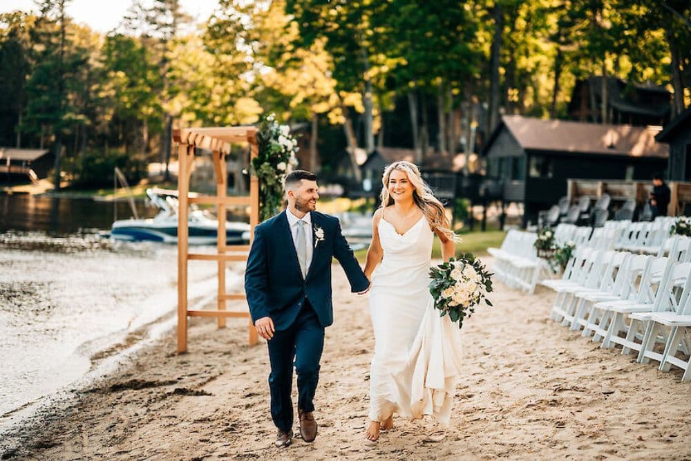 A couple joyfully walks along a beach, celebrating their wedding surrounded by trees and boats.