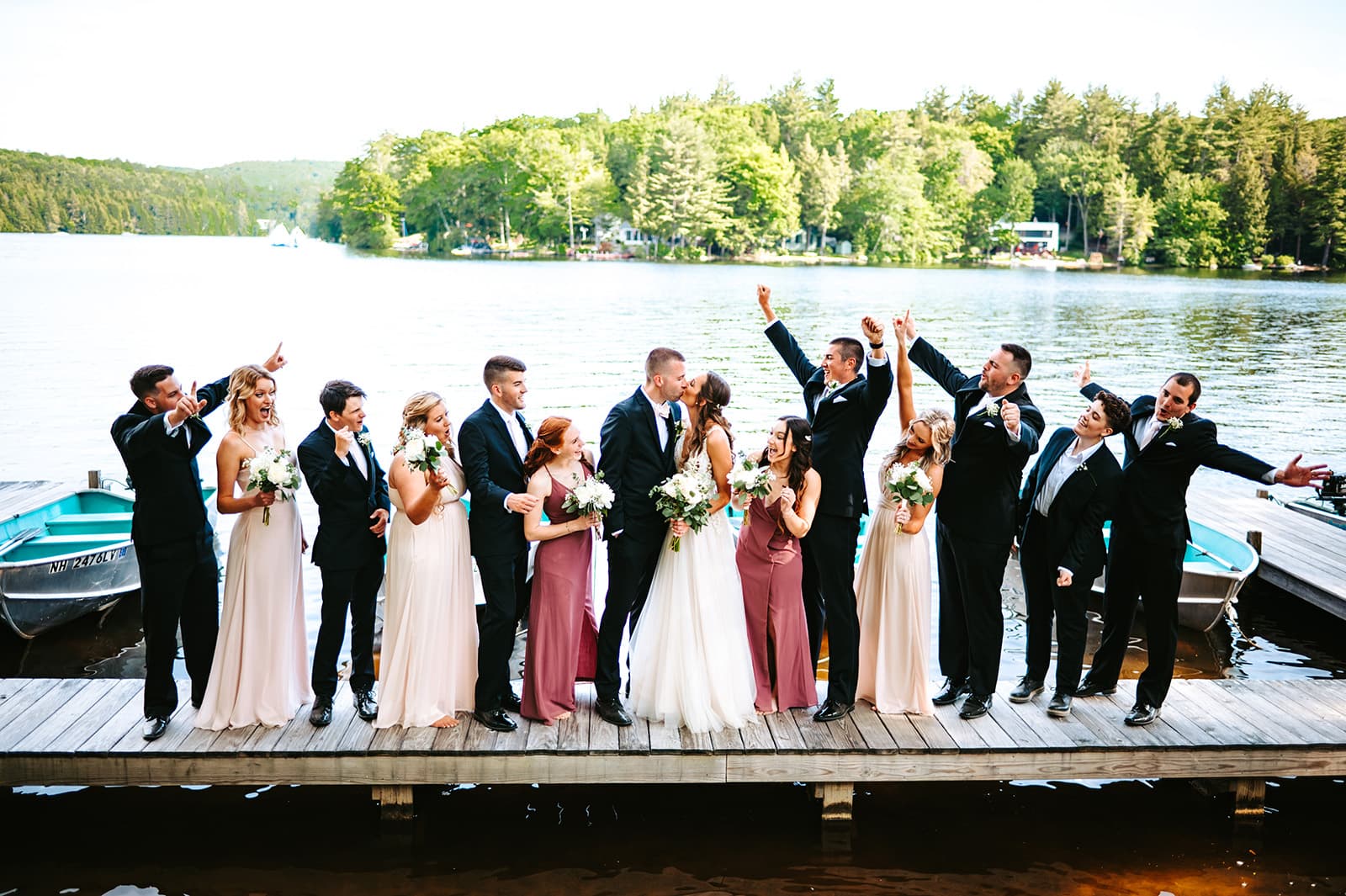 A joyful wedding party poses on a dock by a lake, celebrating with flowers and cheerful gestures.