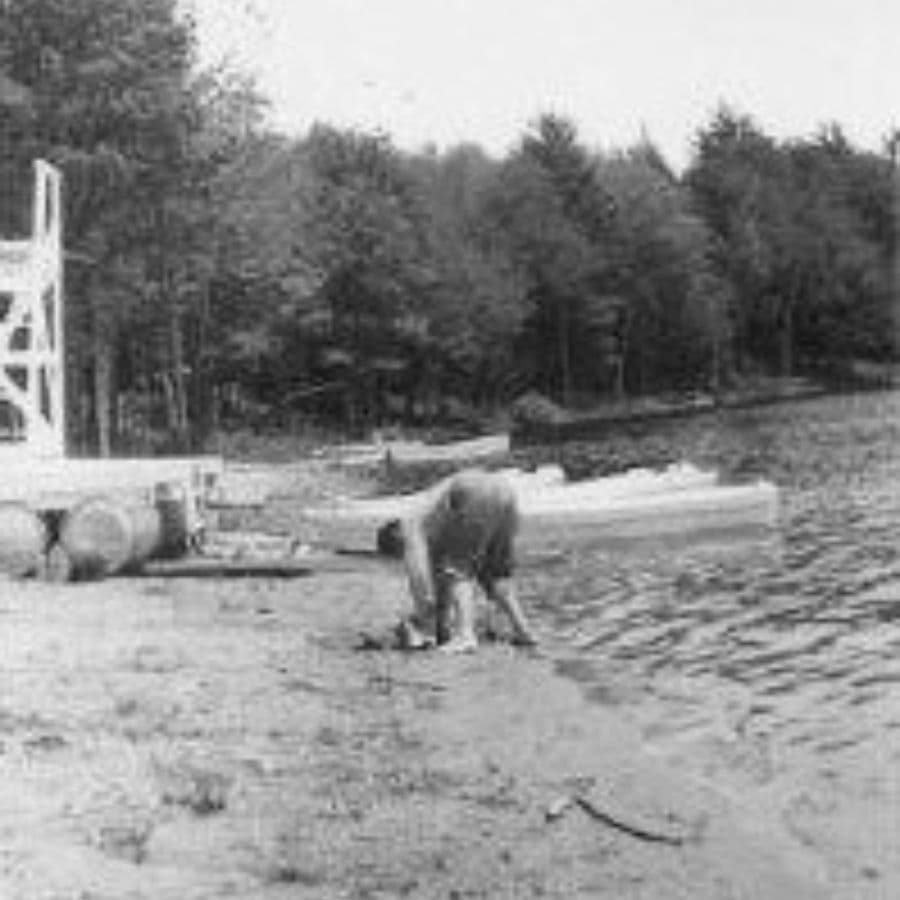 A person bends over near the water's edge, surrounded by trees and canoes.