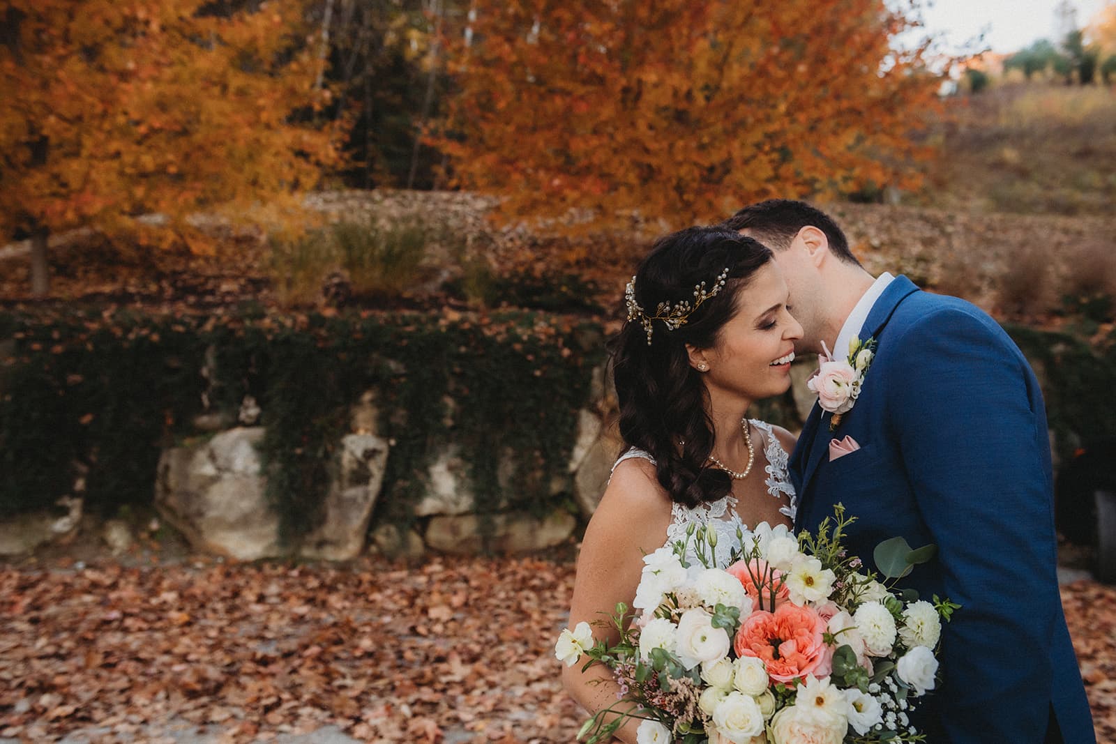 A couple shares a tender moment outdoors in front of autumn foliage, with the bride holding a vibrant bouquet.