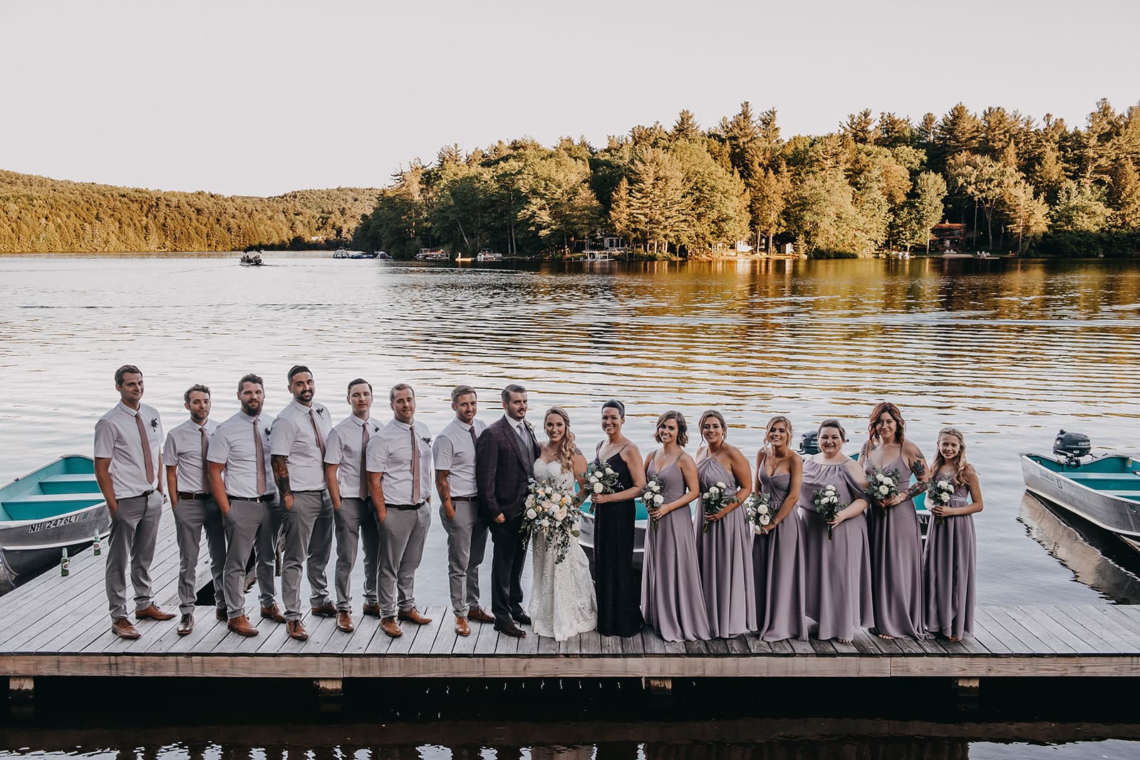 A wedding party poses on a dock by a serene lake surrounded by trees.