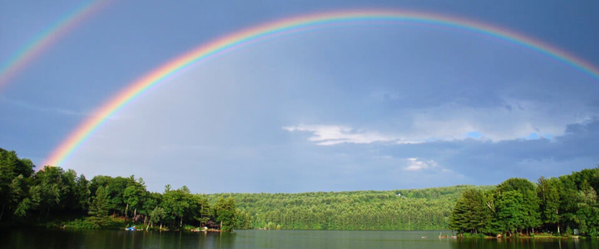 A vibrant double rainbow arcs over a serene lake surrounded by lush green trees.