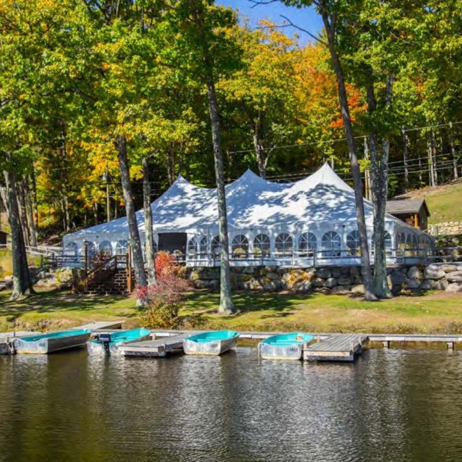 A large white tent is set up near a calm lake surrounded by colorful trees and boats docked nearby.