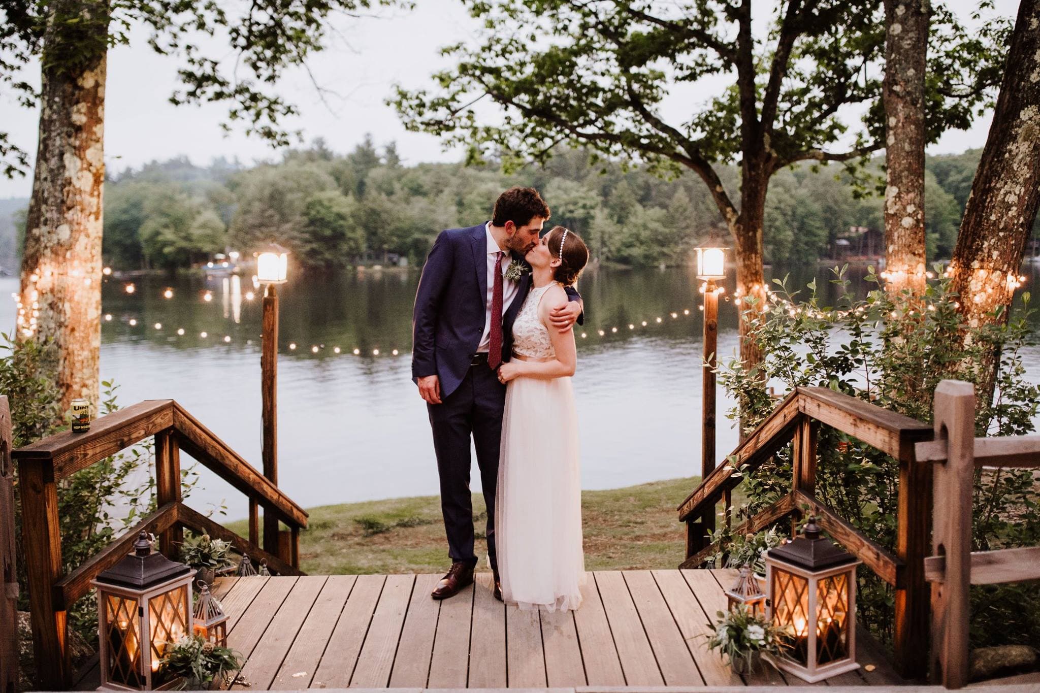 A bride and groom kiss on a wooden deck overlooking a serene lake, surrounded by trees and glowing lanterns.