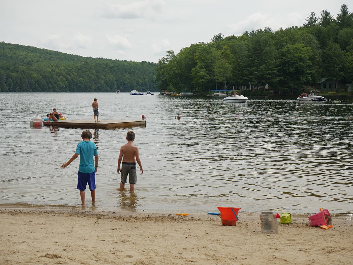 Children play at the lake shore while others swim and enjoy a floating dock in the background.