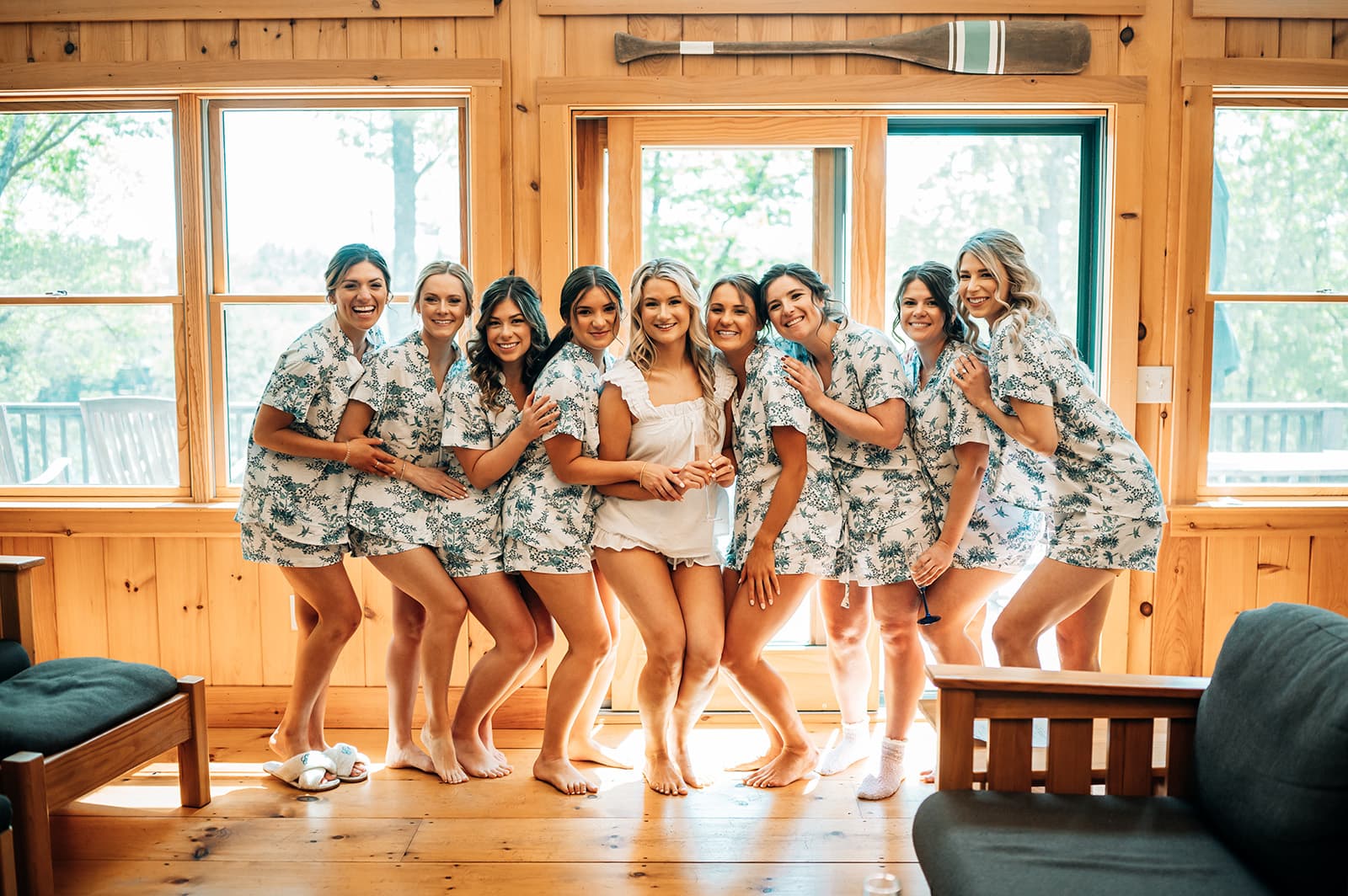 A group of eight women in matching floral pajamas smiles together in a sunlit room with wooden walls and large windows.