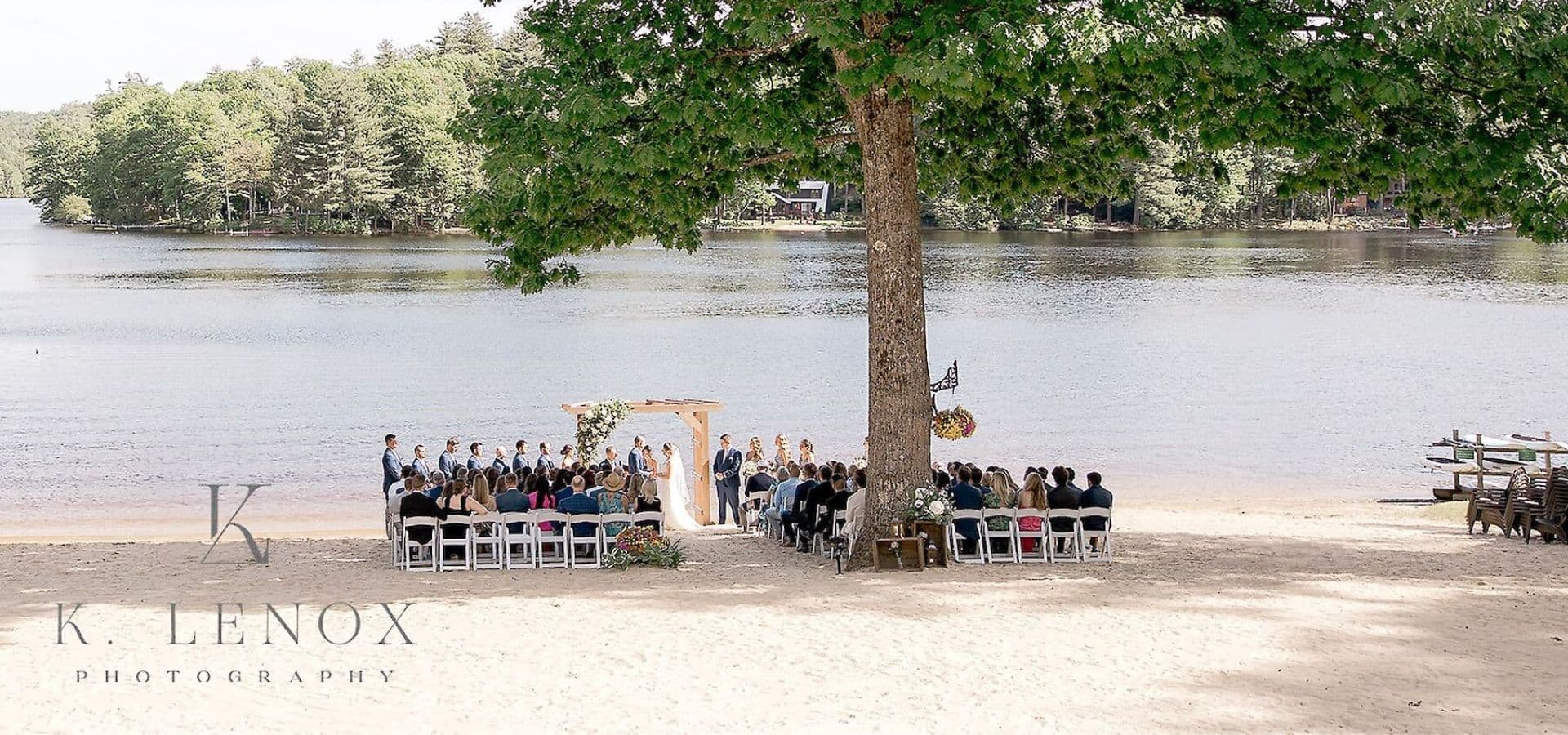A wedding ceremony takes place on the beach by a lake, surrounded by guests seated in rows.