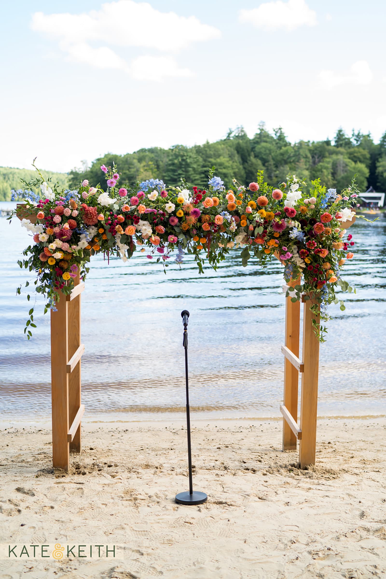 A floral arch with vibrant flowers stands by a tranquil lake, accompanied by a microphone on a sandy beach.