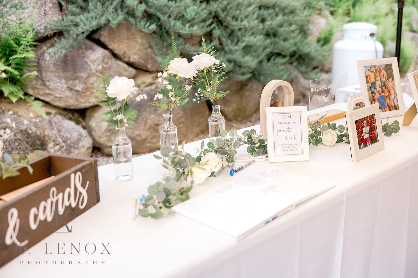 A decorated table featuring flowers, a guest book, and framed photos against a stone backdrop.