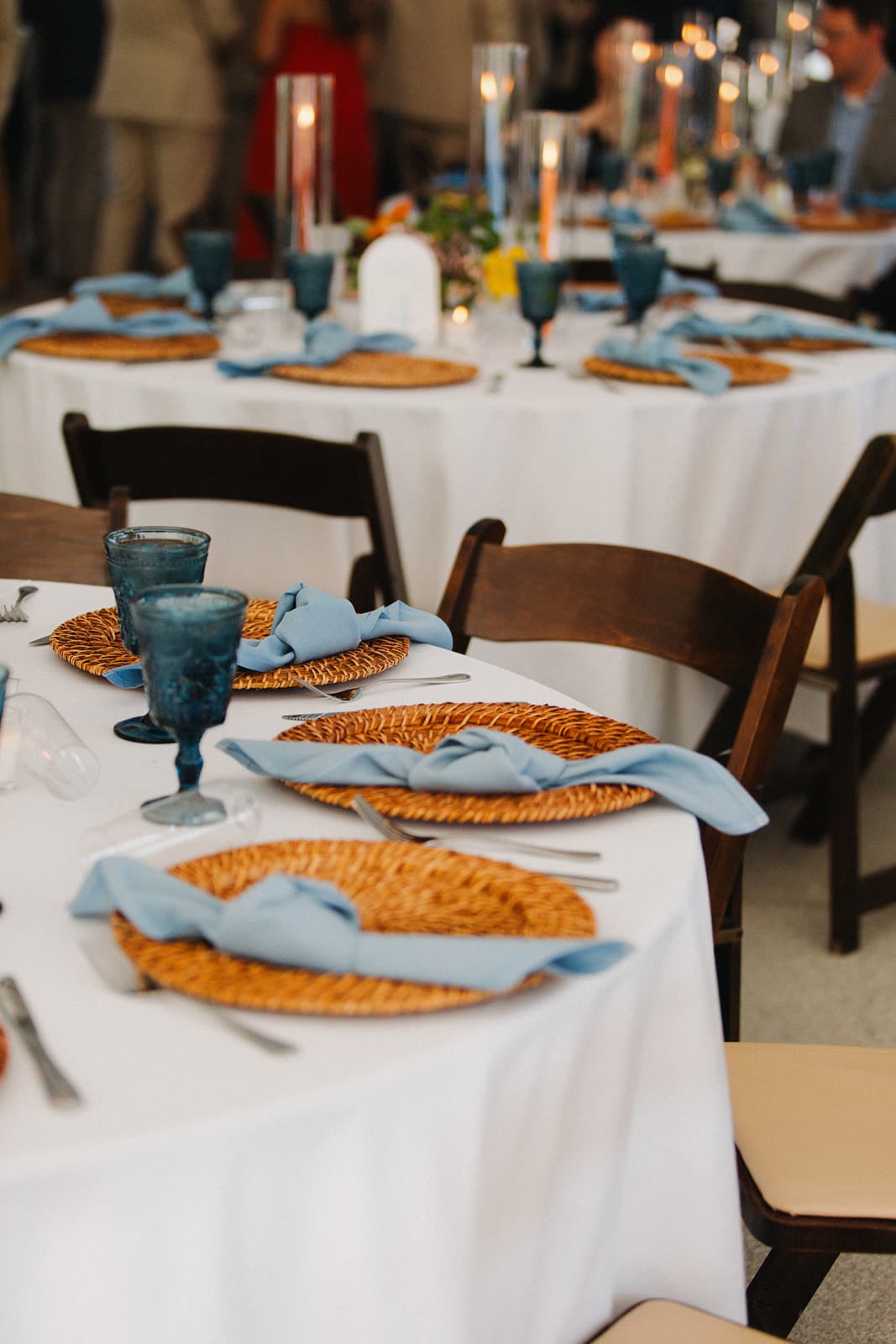 A beautifully set dining table with woven placemats, blue napkins, and glasses, surrounded by a softly lit atmosphere.