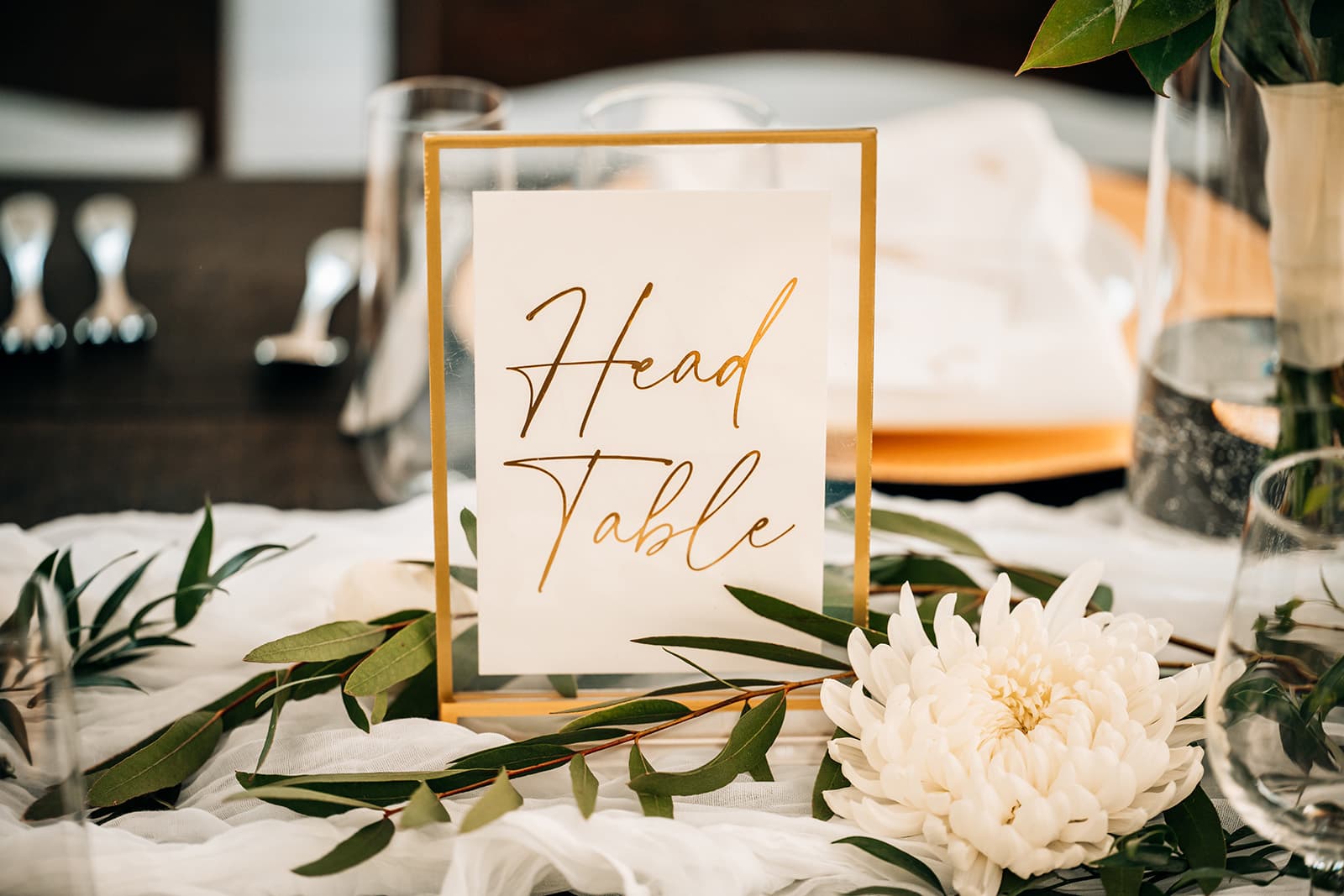 A decorative sign reading "Head Table" surrounded by greenery and a white flower on an elegantly set table.