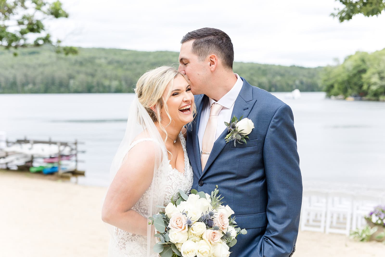 A joyful bride laughs as her groom kisses her forehead by a serene lake.