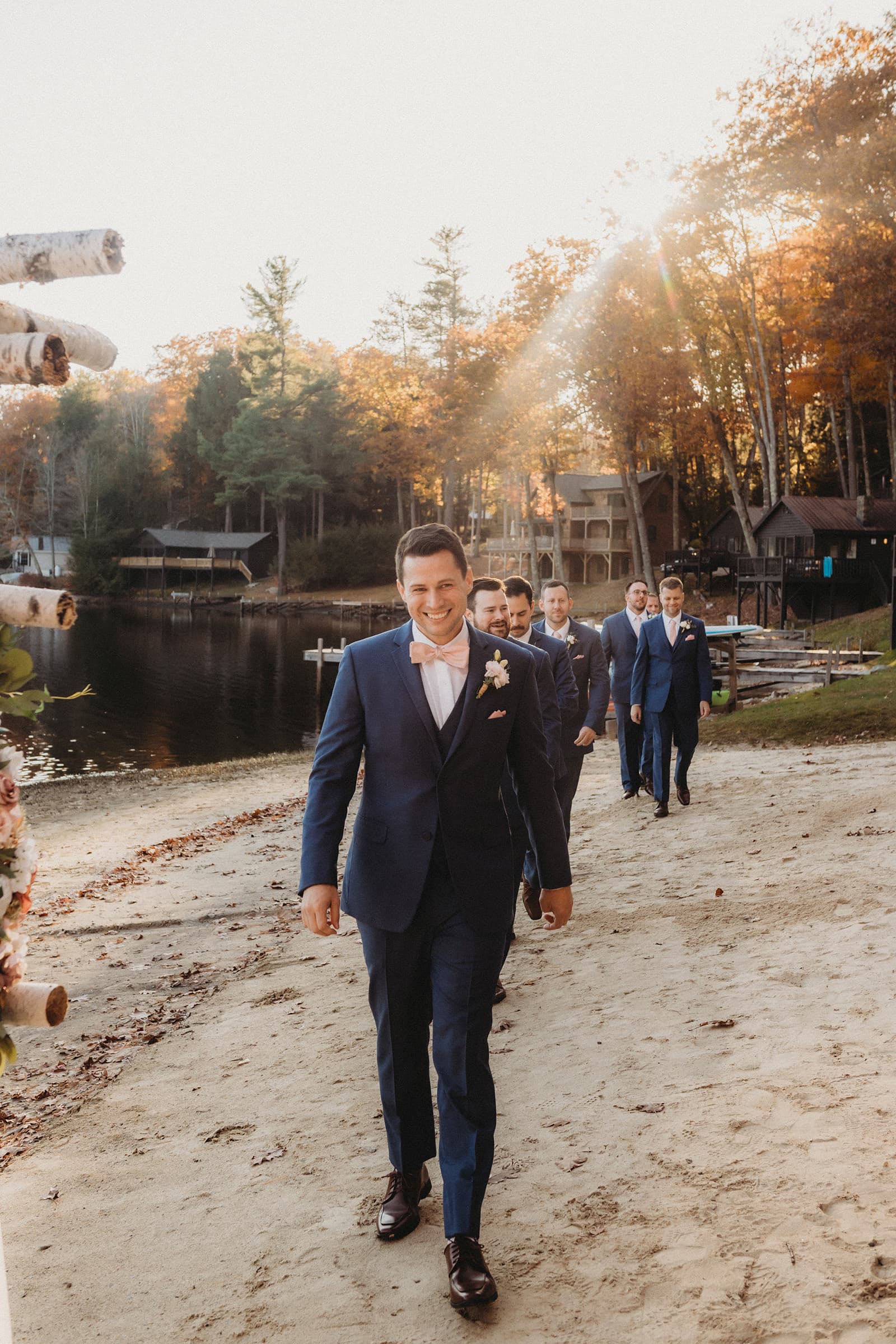 A group of groomsmen walks along a sandy lakeshore in the autumn.
