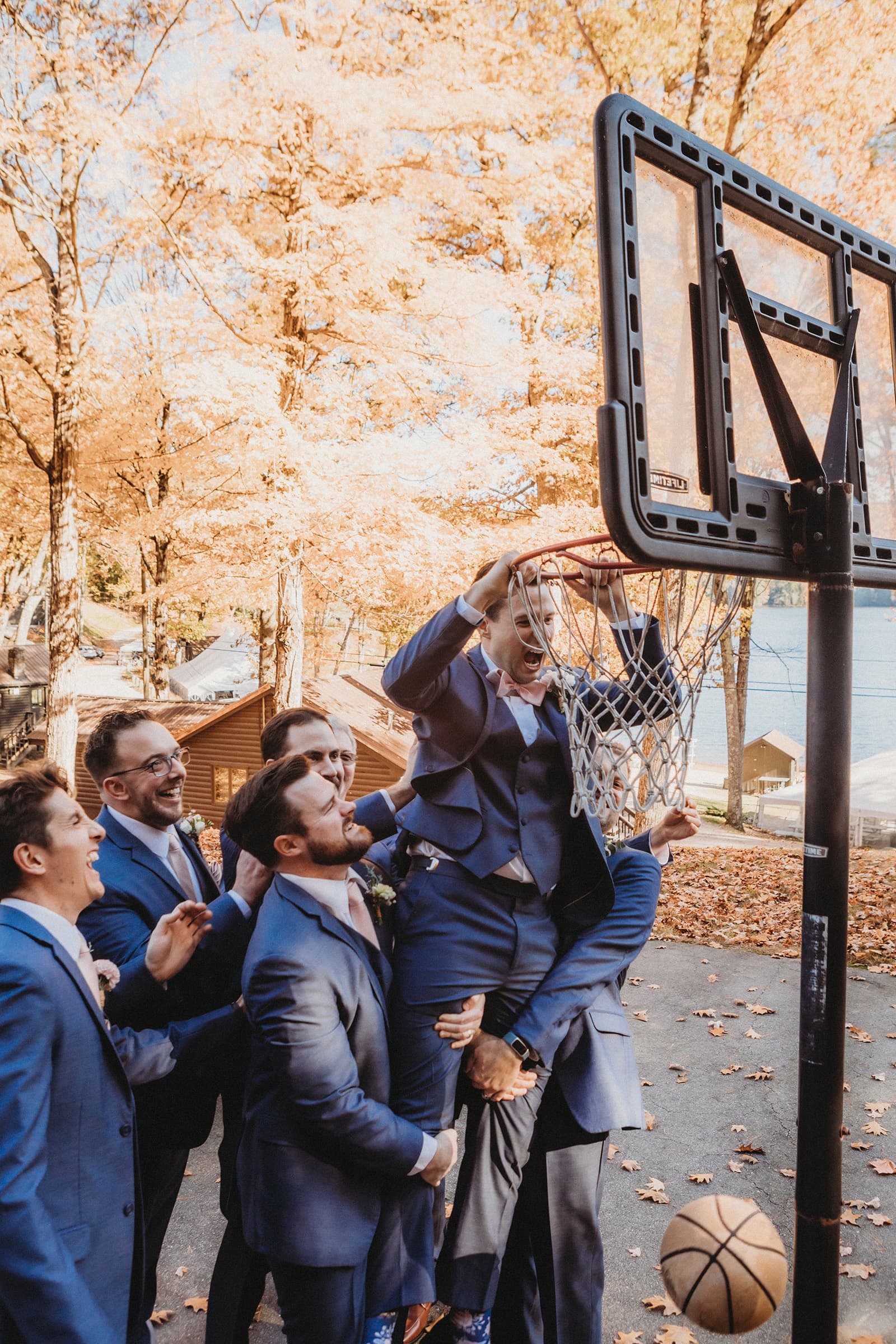 A group of six men in suits celebrates as one jumps and hangs from a basketball hoop surrounded by autumn leaves.