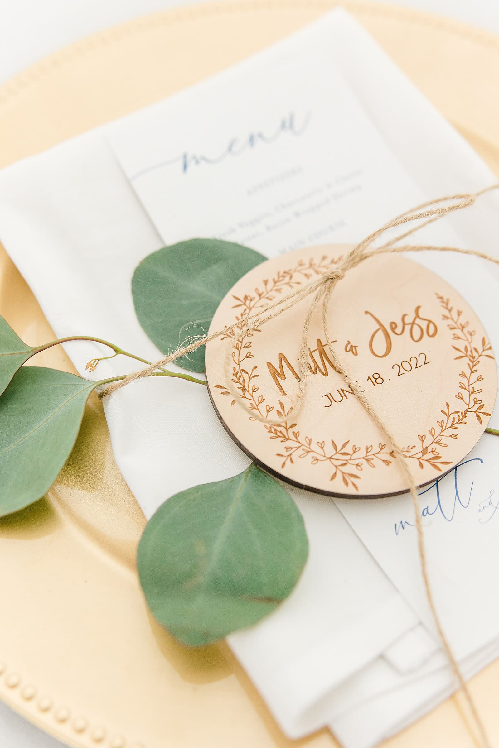 A wooden name tag with a floral design, tied with twine, rests on a folded white napkin on a golden plate.