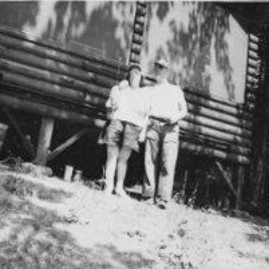 A couple stands together in front of a wooden cabin.