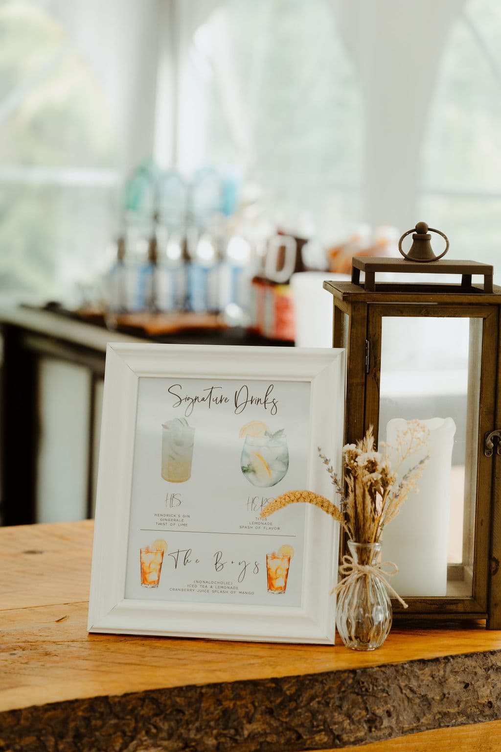 A framed menu displays signature drink options next to a decorative lantern and a small vase of dried flowers on a wooden table.