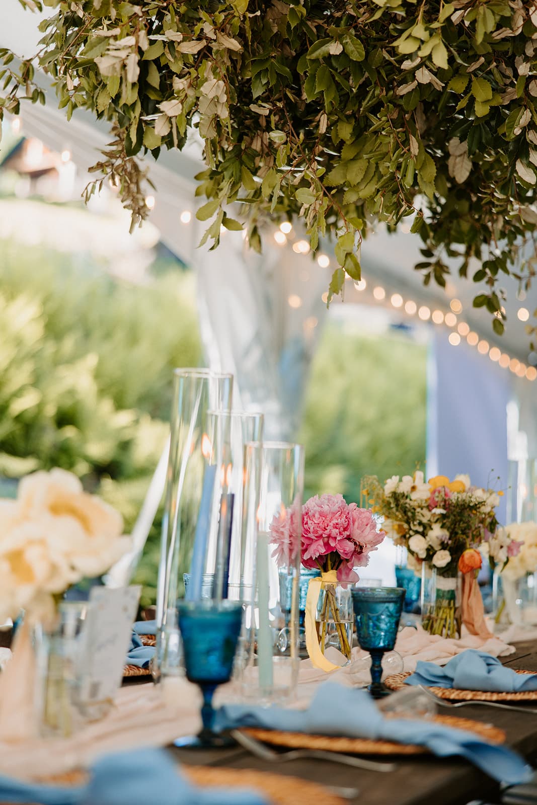A beautifully arranged outdoor table adorned with flowers, candles, and blue glassware under a leafy canopy.