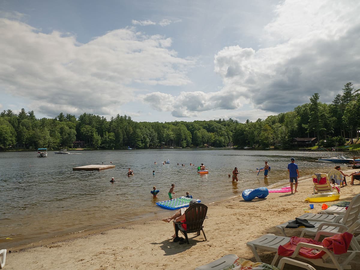 A sunny lakeside scene with people swimming, relaxing on the beach, and a floating dock in the background.