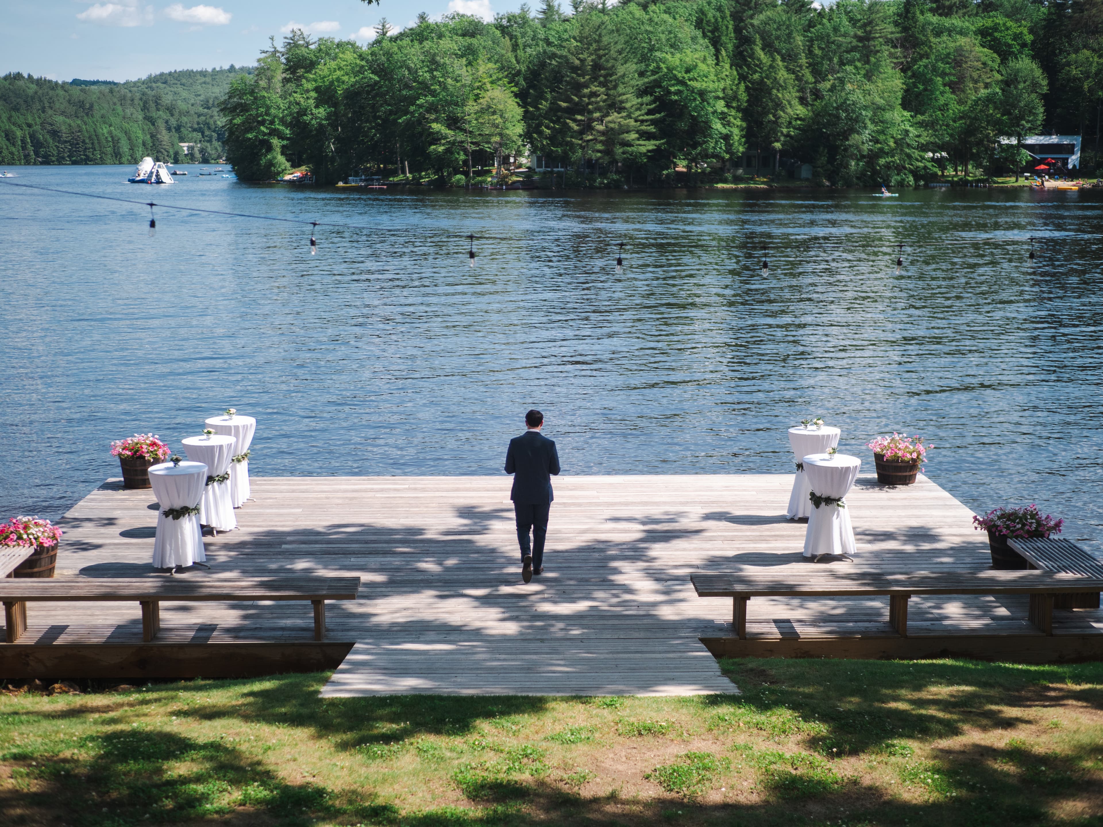 A man stands on a wooden dock overlooking a serene lake with tables set up for an event.