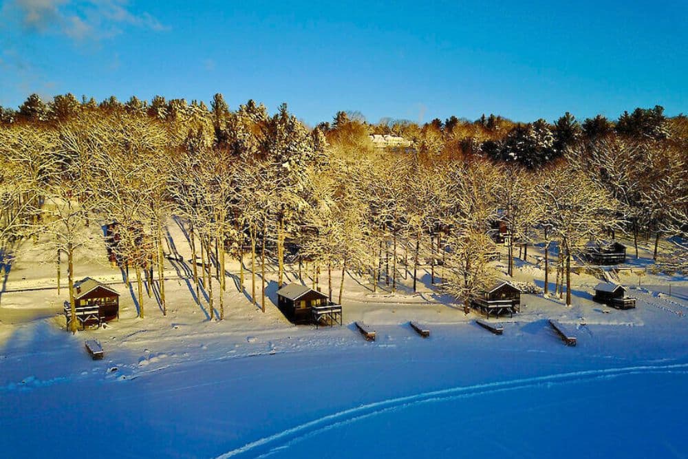 Aerial view of snow-covered cabins beside a frozen lake surrounded by trees.