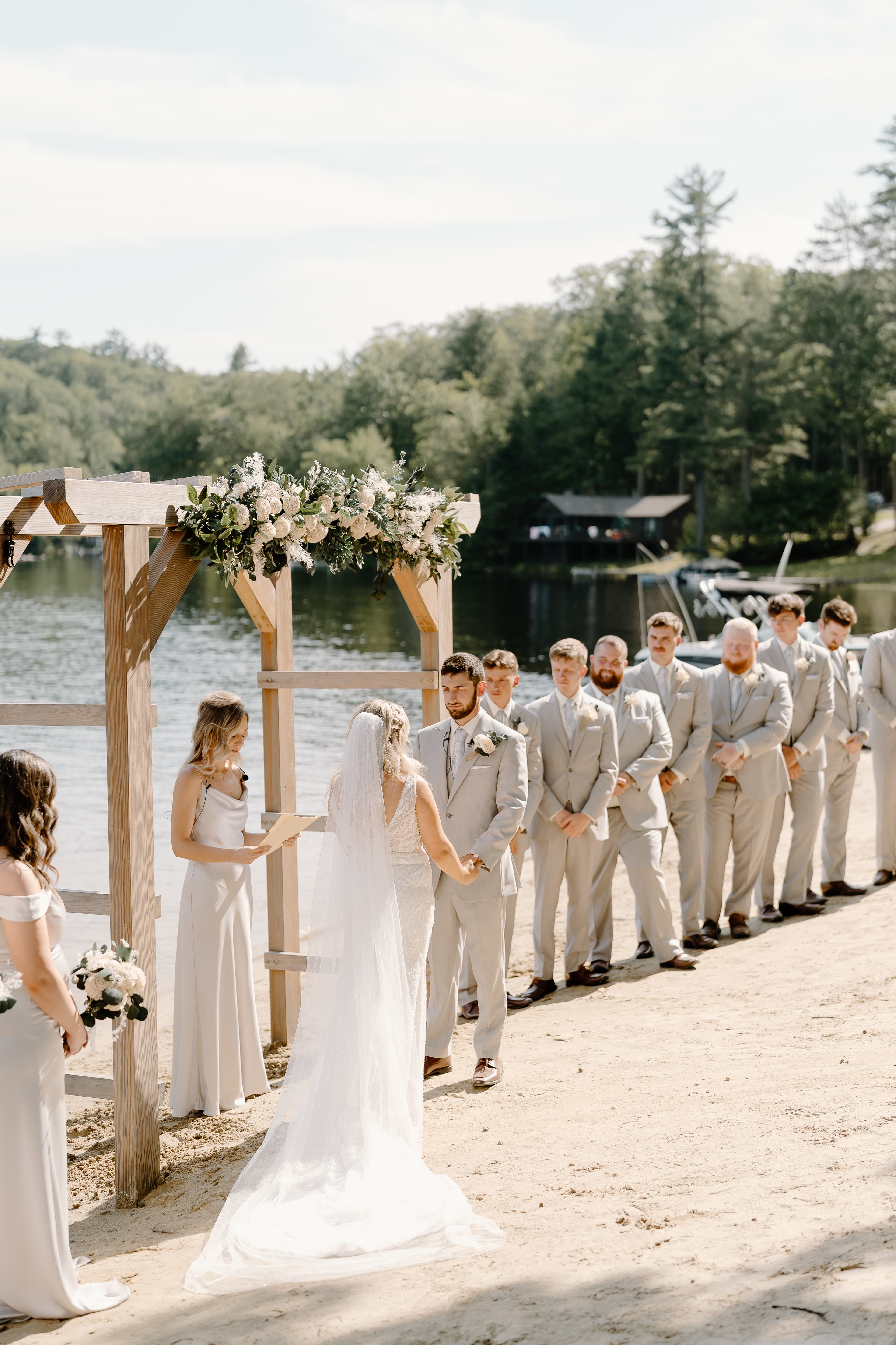 A bride and groom stand under an embellished arch by a lake, surrounded by wedding party members.