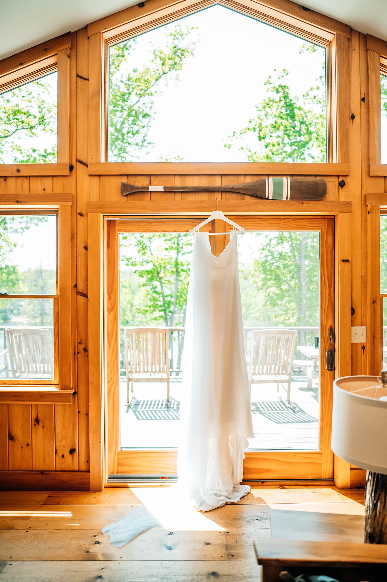 A white wedding dress hangs in front of large windows in a wooden interior.