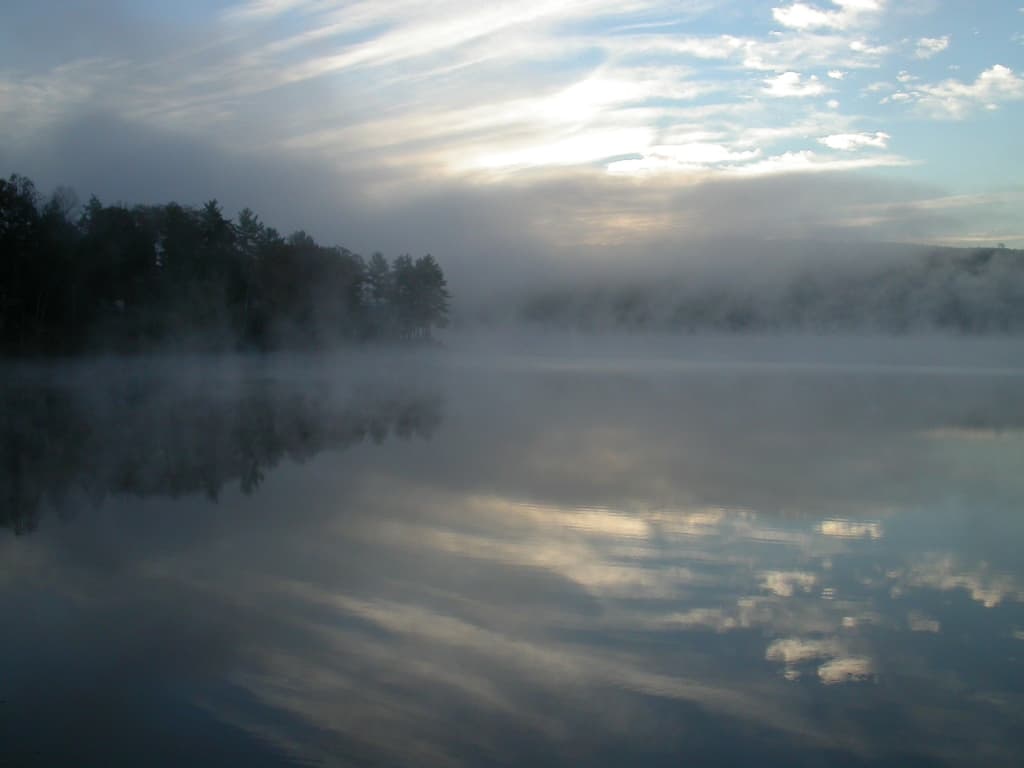 A tranquil lake scene with mist rising and soft clouds reflecting in the water.